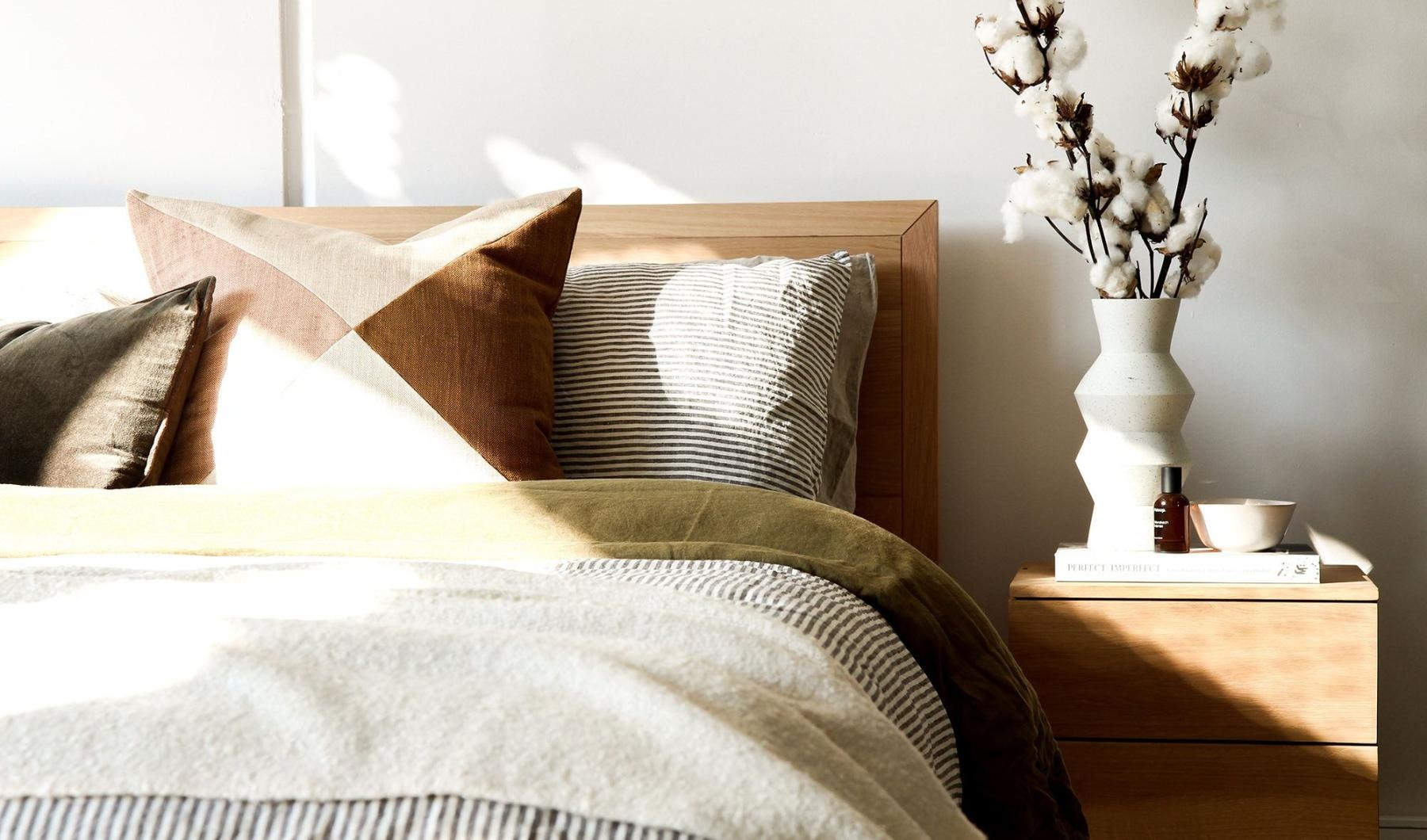swath of sunlight shines across a well made bed and bedside table with a vase of cotton plants
